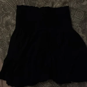 Svart kortkjol med smockad midja - En svart kortkjol med smockad, elastisk midja som sitter snyggt och bekvämt. Kjolen har en lätt utställd form och är perfekt för dig som gillar en enkel men stilren look. Materialet känns mjukt och följsamt.