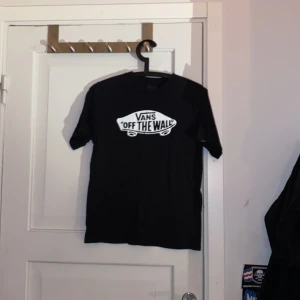 Svart Vans Off The Wall t-shirt - Svart t-shirt från Vans med klassiskt 'Off The Wall'-tryck i vitt på bröstet. Modellen är rak med rund hals och korta ärmar. Perfekt för dig som gillar streetwear och skatekänsla. Materialet är mjuk bomull som är skönt mot huden.