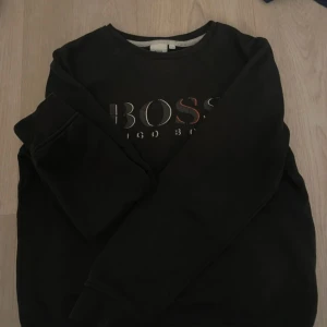 Svart sweatshirt från Hugo Boss - Svart sweatshirt från Hugo Boss med stort BOSS-tryck i vitt och rött framtill. Klassisk rund halsringning och långa ärmar. Mjuk insida och relaxed fit, perfekt för en chill och stilren look.