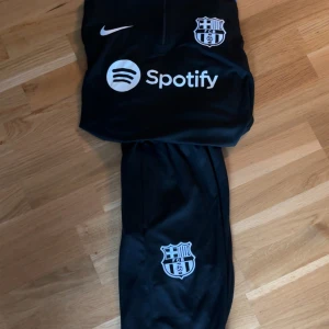 Nike FC Barcelona svart träningströja - Svart långärmad träningströja från Nike x FC Barcelona med halvlång dragkedja, klubbmärke på bröstet och sponsorlogga. Snygga blå och röda detaljer på ärmarna. Perfekt för fotbollsträning eller chill.