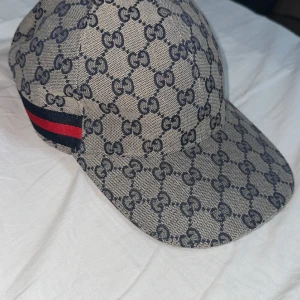 Gucci keps med GG-mönster och röd/blå detalj - Snygg keps från Gucci med klassiskt GG-mönster i mörkblått på beige bakgrund. Kepsen har en röd och blå rand på sidan och justerbar rem i skinn baktill. Materialet är bomull och skinn, perfekt för dig som vill ha en lyxig streetstyle-look.