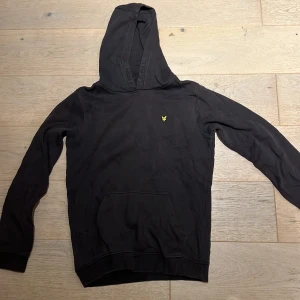 Svart hoodie från Lyle & Scott Junior - Svart hoodie från Lyle & Scott Junior med klassisk gul logga broderad på bröstet. Tröjan har huva, känguruficka och långa ärmar. Tillverkad i mjuk bomull, perfekt för chill dagar eller när du vill ha en enkel och stilren look.