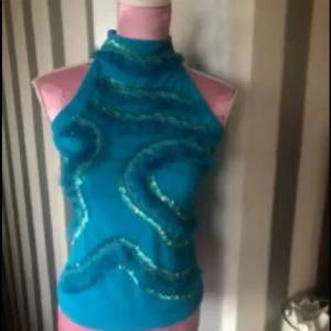 Turkos mesh polotopp med paljetter NY - Säljer en turkos mesh polotopp med transparent material och glittriga paljettdekorationer i vågiga mönster framtill. Toppen har hög hals och är ärmlös med en snygg öppning i nacken. Perfekt för dig som gillar att sticka ut med färg och detaljer.