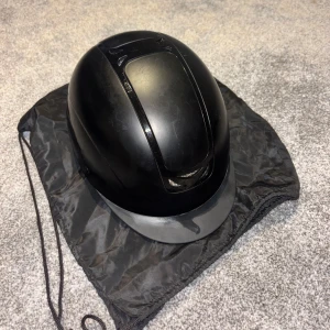 Svart ridhjälm från JH Collection - Säljer en svart ridhjälm från JH Collection med klassisk rundad form och justerbart hakband. Hjälmen har ventiler upptill och kommer med en svart förvaringspåse. Perfekt för dig som rider och vill ha en stilren och säker hjälm. Har aldrig ramlat av med den men den har lite repor från grus, grenar mm.