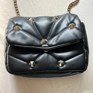 Svart axelväska med nitar - Snygg svart axelväska i skinnimitation med quiltad design och guldfärgade nitar. Väskan har en kedja och läderrem som axelband, samt ett rymligt fack med innerficka. Perfekt för dig som gillar edgy detaljer och stilren look.