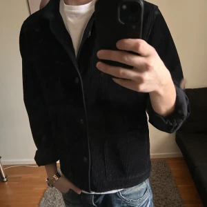 Svart manchester overshirt Dressmann  - Svart overshirt från Dressmann i mjuk manchester. Klassisk rak passform med tre stora fickor framtill och knappar hela vägen. Perfekt att slänga över en t-shirt för en chill och stilren look. Storlek S.