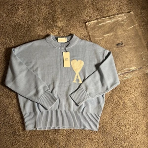 Ljusblå stickad tröja från Ami - Snygg ljusblå stickad tröja från Ami Alexandre Mattiussi med ett stort vitt hjärta och bokstaven A på bröstet. Tröjan har ribbade muddar vid ärmslut och nederkant samt rund hals. Perfekt för dig som gillar stilrena detaljer och mjuka material.