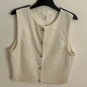 Vit väst med knappar från H&M - Stilren vit väst från H&M med rund halsringning och silverfärgade knappar framtill. Perfekt att styla över en t-shirt eller skjorta för en clean look. Strl XS