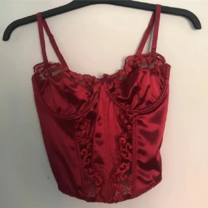 Röd satin korsett med spetsdetaljer - Snygg röd korsettopp i glansig satin med vackra spetsdetaljer längs byst och framtill. Aldrig använd endast testad. stretch i den och går att justera banden oxå💕 vet inte vart den är ifrån men hon jag fick den ifrån sa den va från hukemöller men finns ingen lapp i💕