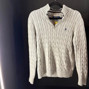 Grå stickad half zip-tröja Polo Ralph Lauren - Kabelstickad grå tröja från Polo Ralph Lauren med half zip och hög krage. Klassisk broderad logga på bröstet i mörkblått. Ribbstickade muddar och krage med randig insida. Perfekt för lager-på-lager under kyliga dagar.