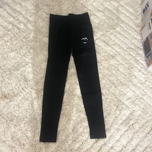 Svarta Peach leggings med scrunch - Säljer ett par svarta leggings från Peach med hög midja och scrunch-detalj bak för extra shaping. Materialet är stretchigt och mjukt, perfekt för träning eller chill. Diskret logga framtill och bak, samt ett litet vitt tryck med smiley på ena benet.