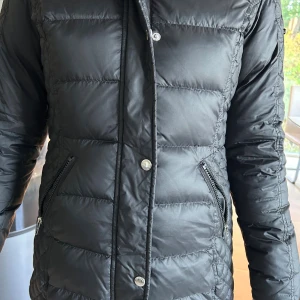 Svart Rockandblue dunjacka med päls - Snygg svart dunjacka från Rockandblue med fluffig svart fuskpäls på huvan. Jackan har quiltad design, dragkedjor på fickor. 