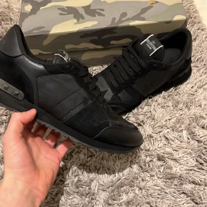 Valentino Garavani svarta sneakers - Svarta Valentino Garavani sneakers med coola nitar på hälen och diskret camomönster. Skorna har snörning, rund tå och är gjorda i en mix av skinn och mocka. Kommer med originalkartong, extra skosnören och tags.