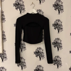 Svart croppad långärmad polotopp - Säljer en svart croppad polotopp med lång ärm och cool öppen detalj över bysten. Toppen har en tight passform och är tillverkad i ett ribbat material som ger en snygg struktur. Perfekt för dig som gillar trendiga och stilrena plagg med en twist.