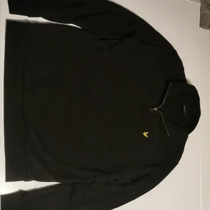 Svart half zip tröja Lyle & Scott - Svart långärmad tröja från Lyle & Scott i storlek L. Tröjan har en snygg half zip-dragkedja vid kragen och den klassiska gula fågel-loggan broderad på bröstet. Tillverkad i mjuk bomull, perfekt för chill dagar. Minimalistisk och stilren design. Helt oanvänd, och ny pris 999kr.