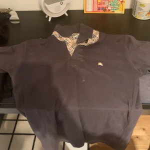 Mörkblå Burberrys London skjorta - Snygg mörkblå skjorta från Burberrys London med klassiskt rutig krage och knappslå i beige, svart och röd. Ikonisk broderad logga på bröstet. Skjortan har korta ärmar och är gjord i mjuk bomull. Perfekt för dig som gillar stilrena och tidlösa plagg.