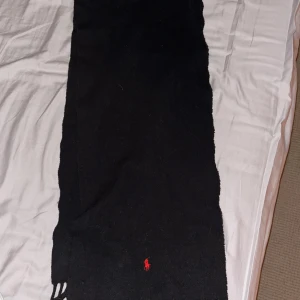 Svart ullhalsduk från Polo Ralph Lauren - Klassisk svart halsduk från Polo Ralph Lauren i mjuk ull med fransar i ändarna. Liten röd broderad logga framtill ger en snygg kontrast. Perfekt accessoar för kalla dagar och enkel att matcha med olika outfits.