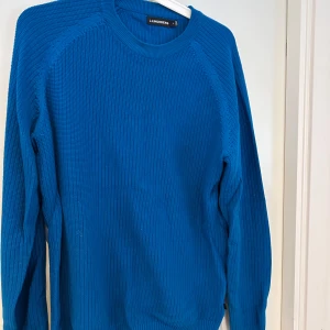 Blå stickad tröja från J.Lindeberg - Snygg blå stickad tröja från J.Lindeberg i storlek M. Tröjan har en ribbad struktur och rund hals, samt långa ärmar. Perfekt för dig som gillar stilrena och klassiska plagg med lite edge. Passar dig som vill ha en enkel men cool look.