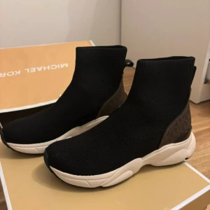 Svarta sock-sneakers från Michael Kors - Snygga svarta sock-sneakers från Michael Kors med chunky vit sula och bruna detaljer med MK-logga på hälen. Ovandelen är stickad och följsam för en modern look. Perfekta för dig som vill ha både stil och komfort i vardagen.aldrig använda dessutom