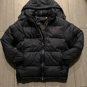 Svart pufferjacka från Polo Ralph Lauren - Svart pufferjacka från Polo Ralph Lauren med huva och röd logga på bröstet. Jackan har flera dragkedjeförsedda fickor, quiltad design och justerbar nederkant. Perfekt för kalla dagar och har en clean, sportig look.