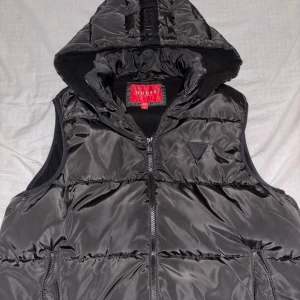 Svart väst med luva från Guess - Svart väst från Guess med mellan storlek på luvan och dragkedja framtill. Västen har ett quiltat mönster och Guess-logga på bröstet samt på luvan. Utsidan är inte så mycket glansig med och insidan är mjuk fleece. Perfekt för lager-på-lager.