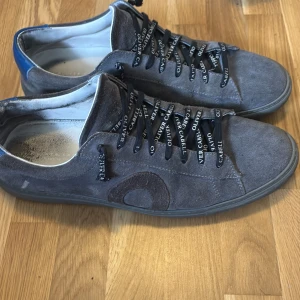 Grå mockasneakers från Oliver Cabell - Snygga grå sneakers från Oliver Cabell i mocka med svarta snören där märket står tryckt i vitt. Skorna har rund tå, platt sula och diskret logga på sidan. Tillverkade i Italien och har en stilren, minimalistisk look som passar till det mesta.