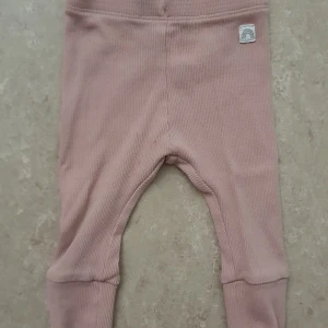 Byxor - Supersöta ribbade babybyxor från Lindex i en mjuk, ljusrosa nyans. Byxorna har breda muddar vid bensluten och en liten tygpatch med regnbågsmotiv framtill. Tillverkade i bomull med lite elastan för extra stretch och komfort.