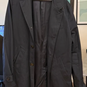 Rock från Riley *M* - Stilren rock från Riley med klassisk passform och två knappar framtill. Rocken har snedställda fickor, knappdetaljer vid ärmslut och slät baksida. Fodret är glansigt och ger en lyxig känsla. Perfekt för dig som gillar en enkel och elegant look.