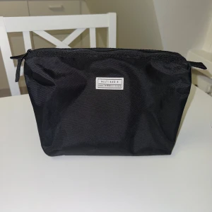 Necessär - Säljer en svart necessär. Necessären har en enkel, stilren design med dragkedja upptill och en liten vit etikett framtill. Tillverkad i slitstark polyester.