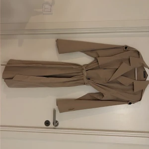 Beige trenchcoat - Snygg beige trenchcoat från Shein med klassisk krage, långa ärmar och bälte i midjan. Jackan har stilrena detaljer som axelklaffar och knappar vid ärmslut. Perfekt för dig som vill ha en clean och tidlös look.
