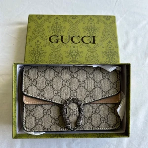 Gucci beige axelväska med kedja - Snygg beige axelväska  Gucci med klassiskt GG-mönster och silverfärgad kedja. Väskan har en dekorativ spänne framtill och insida i ljusbrunt skinn. Perfekt storlek för det viktigaste och riktigt lyxig vibe.