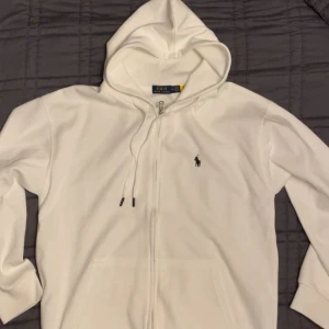 Vit hoodie från Polo Ralph Lauren (prisförslag) - Vit hoodie från Polo Ralph Lauren i storlek XL. Sitter som en L. Tröjan har huva med snörning, dragkedja framtill och klassisk Polo-logga broderad på bröstet. Mjukt material och ribbade muddar vid ärmslut och nederkant. Perfekt för chill dagar.