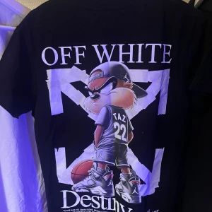 Off white tshirt - Säljer en svart t-shirt från Off-White med stor grafisk print på ryggen av Taz i basketkläder och sneakers, samt Off-White-logga fram. T-shirten har korta ärmar och är gjord i mjuk bomull. Perfekt för dig som gillar streetwear och coola motiv.
