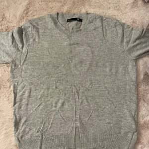 Grå stickad kortärmad tröja - En enkel och stilren grå stickad tröja från Lager med korta ärmar och rund hals. Perfekt för lager-på-lager och passar till både jeans och kjol. Mjuk och skön kvalitet med ribbad kant vid hals, ärmslut och nederkant.