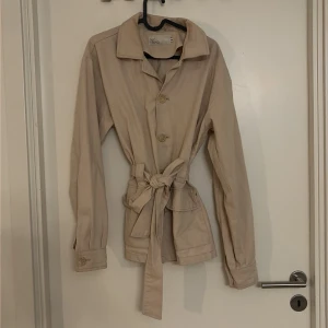 Beige overshirt från NLY Trend - Snygg beige overshirt från NLY Trend med klassisk krage, knappar framtill och ett matchande bälte i midjan. Jackan har långa ärmar och två stora fickor framtill. Perfekt för lager-på-lager och chill stil.