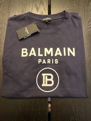 Mörkblå Balmain Paris t-shirt - Snygg mörkblå t-shirt från Balmain Paris med vit logga och tryck framtill. Klassisk rund hals och korta ärmar. Perfekt för dig som vill ha en stilren och trendig look med exklusiv känsla. Tillverkad i mjuk bomull för skön komfort.