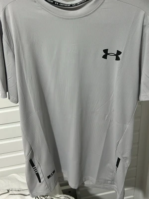Ljusgrå träningströja Under Armour - Ljusgrå kortärmad träningströja från Under Armour med svart logga på bröstet. Tillverkad i ett lätt och ventilerande funktionsmaterial, perfekt för gym eller löpning. Diskreta svarta detaljer vid sidorna ger en sportig look.