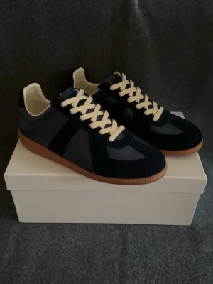 Mörkblå Maison Margiela Gats - Snygga mörkblå Maison Margiela Gats med vit snörning och kontrasterande brun gummisula. Ovandelen är i en mix av mocka och slätt läder, och insidan är ljus. Klassisk siluett som passar till många stilar.