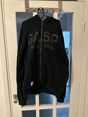 Svart hoodie från GASP Industries - Svart zip-hoodie från GASP Industries med stor logga framtill och tryck på huvan. Hoodien har dragkedja, kängurufickor och ribbade muddar. Tillverkad i mjukt sweatshirtmaterial, perfekt för en avslappnad och sportig stil.