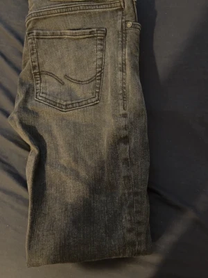 Svarta jeans från Jack and Jones  - Säljer ett par svarta jeans från Jack and Jones  med klassisk femficksmodell och raka ben. Jeansen har en diskret tvättad look och är tillverkade i mjukt denimtyg med stretch för extra komfort. Perfekta till en avslappnad stil.