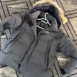 Grå dunjacka från Canada Goose - Snygg grå dunjacka från Canada Goose med stor huva och fuskpälsdetalj. Jackan har flera dragkedjeförsedda fickor och kraftig vaddering som håller dig varm under kalla vinterdagar. Klassisk design med hög krage och justerbar huva.