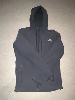 Svart vindjacka från The North Face - Snygg svart vindjacka från The North Face med huva och dragkedjor framtill samt på fickorna. Jackan har lång ärm och logga på bröstet och ryggen. Perfekt för blåsiga dagar och enkel att matcha med olika outfits.