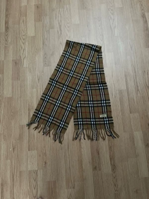 Klassisk beige rutig halsduk Burberry - Säljer en ikonisk beige halsduk från Burberry med det klassiska rutmönstret i svart, vitt och rött. Halsduken har fransar i ändarna och är tillverkad i mjuk ull. Perfekt accessoar för att lyfta din outfit med en tidlös och lyxig känsla. Pris går att diskuteras via snabb affär. Nypris är ca 5000 kronor