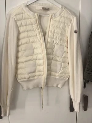 Vit cardiganjacka från Moncler - Snygg vit cardiganjacka från Moncler med quiltad front och stickade ärmar. Dragkedja framtill,  muddar och Moncler-logga på ärmen. Perfekt för lager på lager och ger en clean look. Materialmix av dun och stickat tyg.