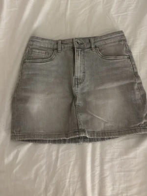 Grå jeanskjol från Kappahl - Säljer en snygg grå jeanskjol från Kappahl i klassisk femficksmodell. Kjolen är kort och har knapp och dragkedja framtill. Perfekt till en avslappnad och trendig look.
