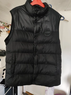 Svart dunväst från Hugo - Snygg svart dunväst från Hugo med hög krage och tryckknappar framtill. Västen har en quiltad design och två fickor på sidorna. Perfekt för lager-på-lager och streetwear-stil. Materialet är glansigt och ger en modern look. Bakvänd kollektion en knapp är borta är mer åt xl än xxl  