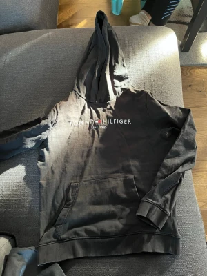 Svart hoodie från Tommy Hilfiger storlek 146/150 - Svart hoodie från Tommy Hilfiger med broderad logga och text 'EST. 1985' på bröstet. Klassisk känguruficka framtill och ribbade muddar vid ärmslut och nederkant. Perfekt för en avslappnad och stilren look. Lite solblekt. Använt skick.