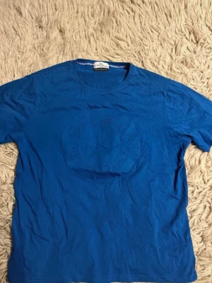 Blå Stone Island t-shirt - Snygg blå t-shirt från Stone Island med diskret broderad logga på bröstet. Klassisk rund hals och korta ärmar. Perfekt till jeans eller shorts för en clean och stilren look. Jag vet inte om den är äkta.