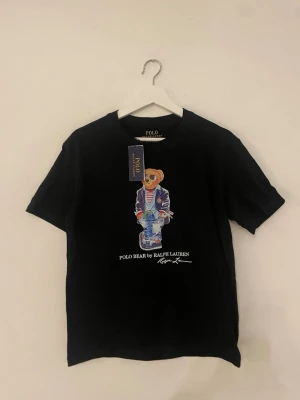 Svart Polo Bear t-shirt från Ralph Lauren - Snygg svart t-shirt från Ralph Lauren med Polo Bear-tryck på framsidan. Klassisk rund hals och korta ärmar. Materialet är mjuk bomull och passformen är normal. Perfekt för dig som gillar streetstyle och ikoniska prints.