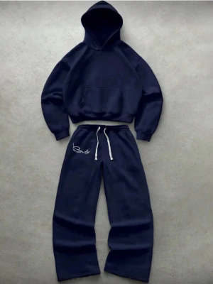 Mörkblå mjukisbyxor med brodyr - Mörkblå mjukisbyxor med vit dragsko i midjan och broderad text på ena benet. Byxorna har avslappnad passform och är gjorda i mjukt, tjockt material som känns riktigt skönt. Perfekta för chill dagar eller när du vill vara bekväm.